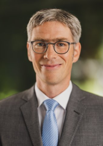 Andreas Honikel-Günther, mit grauer glatter Kurzhaarfrisur, markanten Augenbrauen und ohne Bart. Er trägt eine runde Brille mit dunklem Gestell. Er ist in grauem Jackett mit weißem Hemd und hellblauer Krawatte gekleidet.